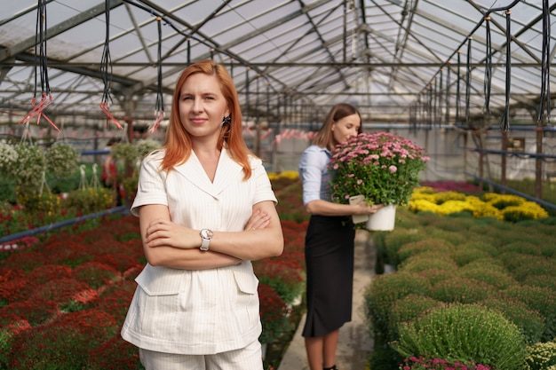 smiling-greenhouse-owner-posing-with-folded-arms-having-many-flowers-colleague-holding-pot-with-pink-chrysanthemums-glass-roof_158595-7031-7352184